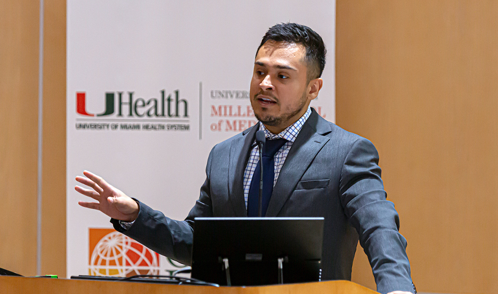 Man in suit speaking at a podium with UHealth and Miller School of Medicine banner