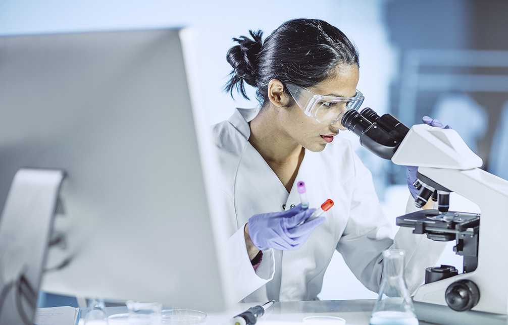 A lab technician in a white lab coat examines a microscope