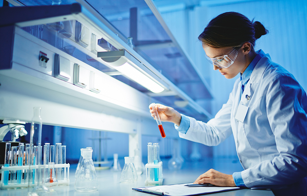A lab technician in a white coat holds up a test tube while she examines her notes