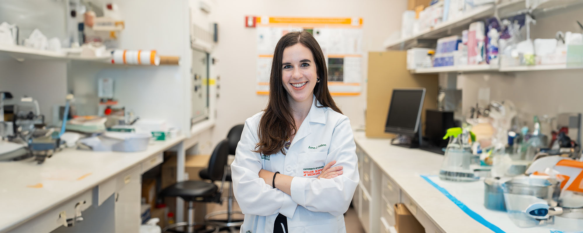 Medical student Anna Hudson, standing with her arms crossed in a laboratory, wearing a white medical coat