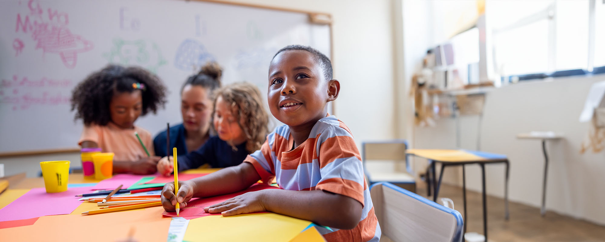 A young boy drawing in a classroom, with other students and a white board in the background