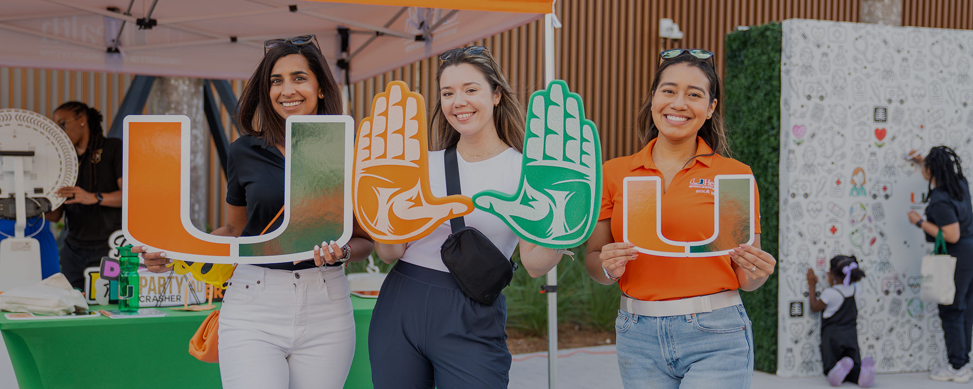 Three women at the UHealth SoLé Mia community event, two holding U signs and one holding two foam hands