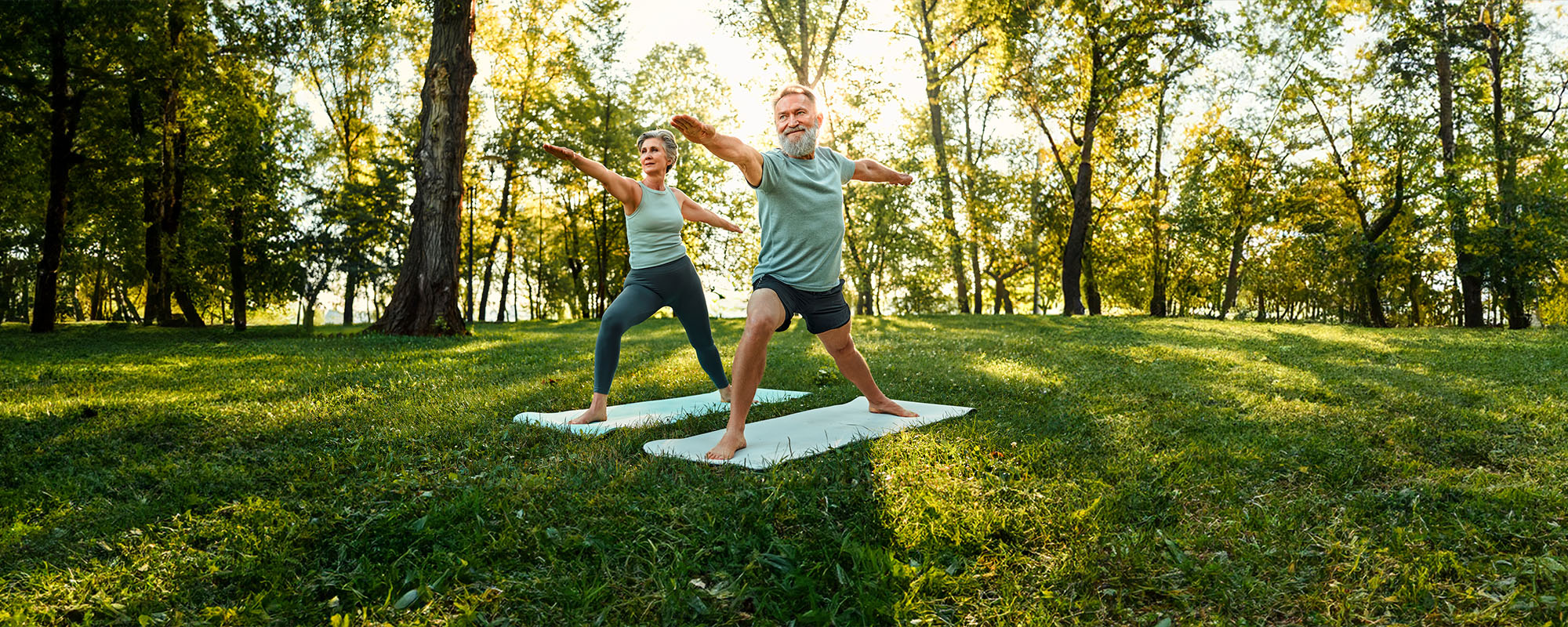 An older man and woman doing yoga on grass, with large trees in the background